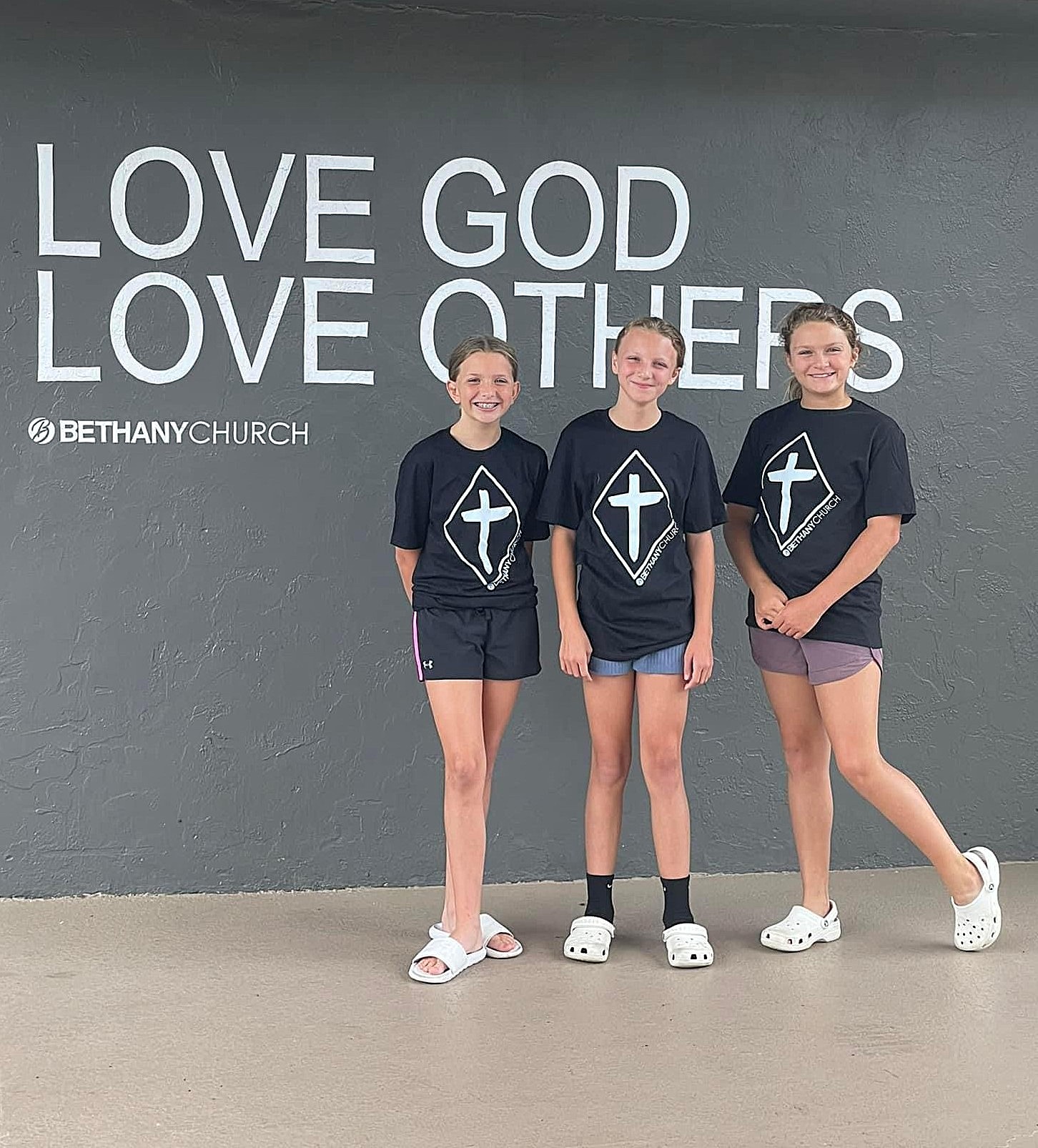 Three Bethany Christian School students standing in front of a wall with the words 'Love God Love Others' at Bethany Church.